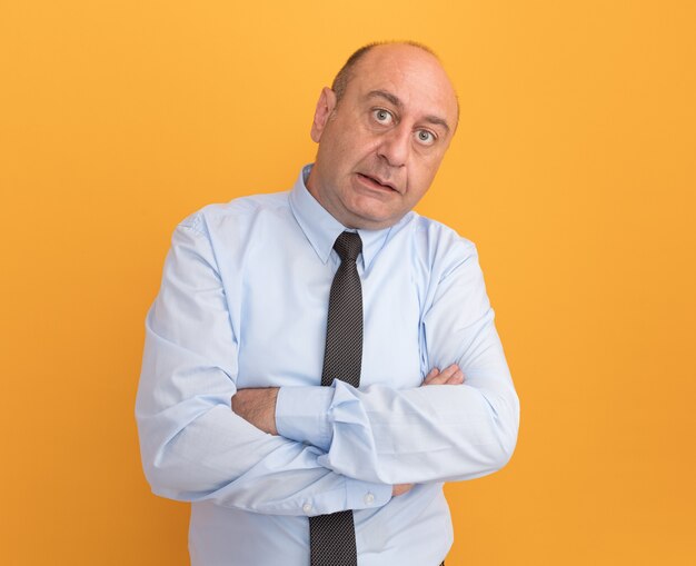 Thinking middle-aged man wearing white t-shirt with tie crossing hands isolated on orange wall