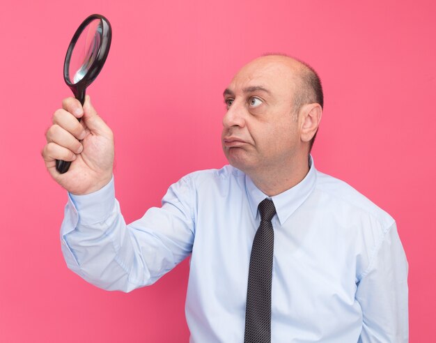 Thinking middle-aged man wearing white t-shirt with tie raising and looking at magnifier isolated on pink wall