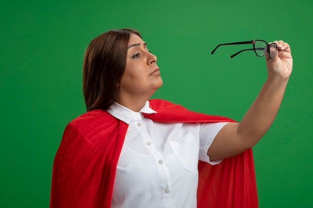 Thinking middle-aged superhero female holding and looking at glasses isolated on green background