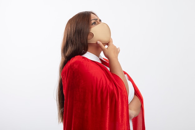 Thinking middle-aged superhero female standing in profile view wearing medical mask putting hand