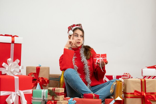 thinking party girl with santa hat holding present sitting around presents on white