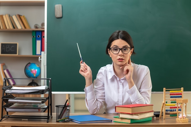 Думая, положив палец на щеку, молодая учительница в очках указывает на доску с указательной палкой, сидя за столом со школьными инструментами в классе