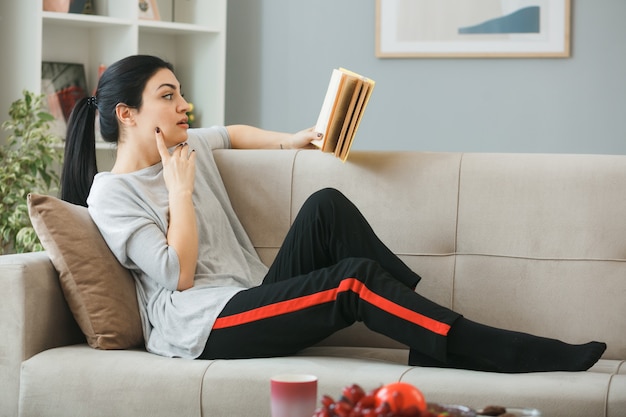 Thinking putting finger on cheek young girl reading book lying on sofa behind coffee table in living room