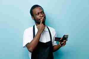 Free photo thinking putting finger on mouth holding card with hair clippers young african american barber in uniform isolated on blue background