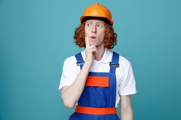 Thinking putting fingers on cheek young builder man in uniform isolated on blue background