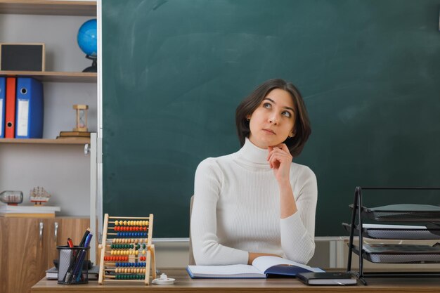 教室で学校の道具を持って机に座っている頬の若い女教師に手を置くことを考えて