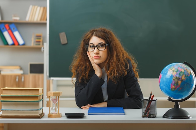 教室で学校の道具を使って机に座って眼鏡をかけている若い女性教師の頬に手を置くことを考えています