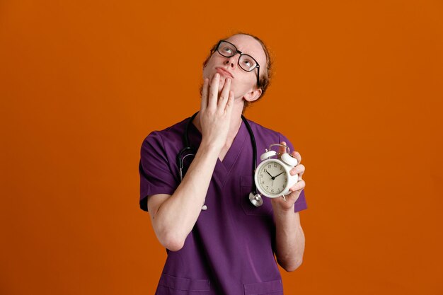 Thinking putting hand on chin holding alarm clock young male doctor wearing uniform with stethoscope isolated on orange background