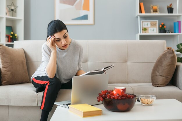 Thinking putting hand on forehead young girl used laptop holding notebook sitting on sofa behind coffee table in living room