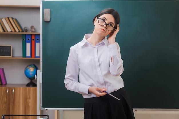 교실에서 칠판 앞에 서 있는 포인터를 들고 머리에 손을 대고 생각하는 젊은 여성 교사