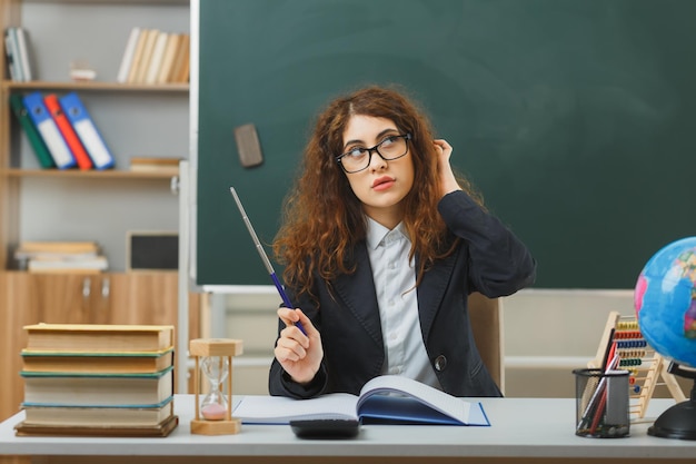 Pensare mettendo la mano sulla testa giovane insegnante femminile che indossa occhiali tenendo il puntatore seduto alla scrivania con strumenti scolastici in classe