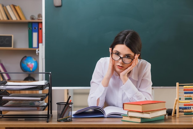 thinking putting hands on cheek young female teacher wearing glasses sitting at desk with school tools in classroom