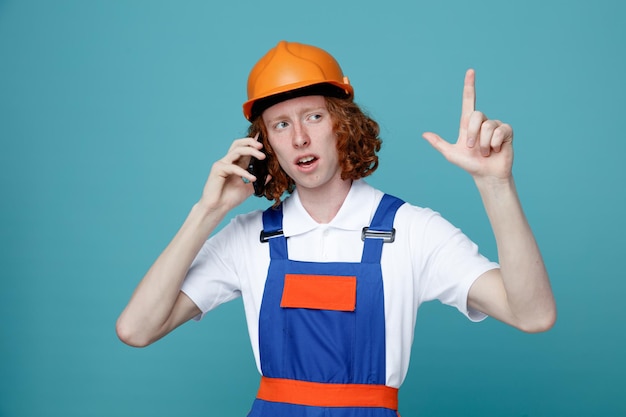 Pensiero che mostra gesto giovane costruttore uomo in uniforme parlare al telefono isolato su sfondo blu