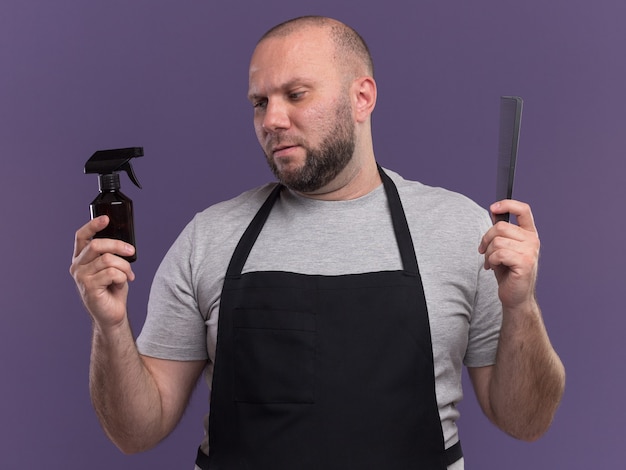 Thinking slavic middle-aged male barber in uniform holding comb and looking spray bottle in his hand isolated on purple wall
