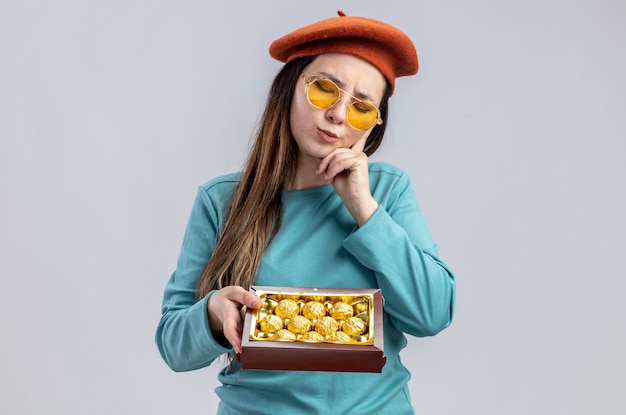 Thinking with closed eyes young girl on valentines day wearing hat with glasses holding box of candies putting hand on cheek isolated on white background