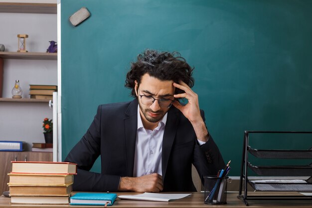 Thinking with lowered head male teacher wearing glasses sitting at table with school tools in classroom