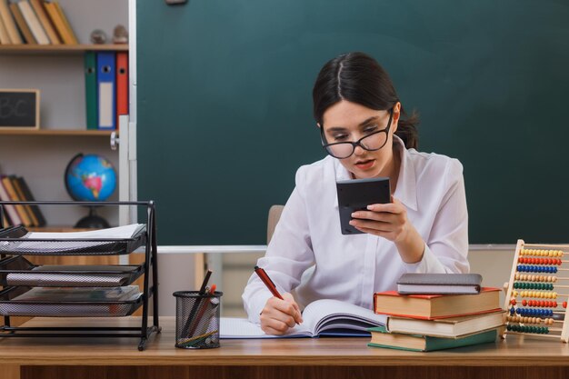 thinking writting young female teacher wearing glasses holding calculator sitting at desk with school tools in classroom