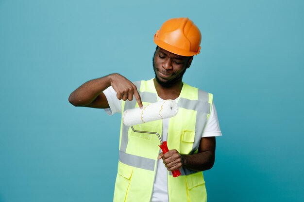 Thinking young african american builder in uniform holding and points at roller brush isolated on blue background