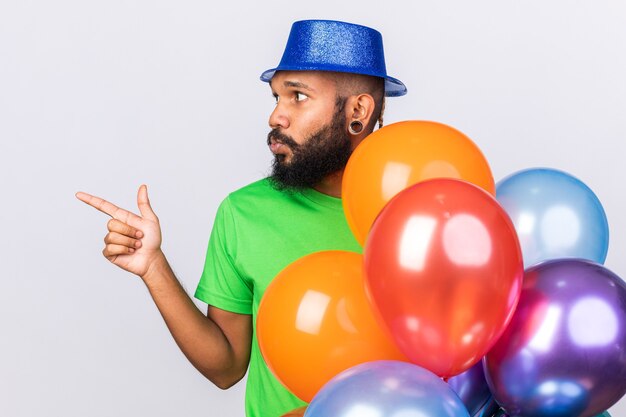 Думающий молодой афро-американский парень в партийной шляпе, стоящий за воздушными шарами, указывает сбоку