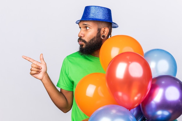 Thinking young afro-american guy wearing party hat standing behind balloons points at side 