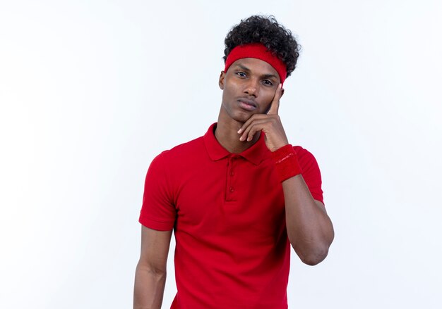 Thinking young afro-american sporty man wearing headband and wristband putting finger on face isolated on white