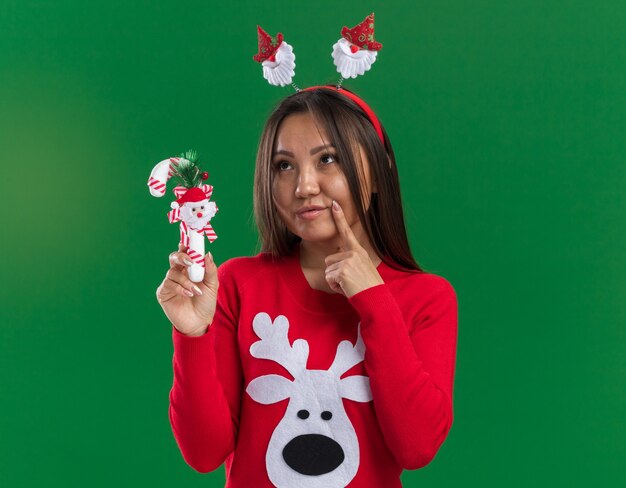 Thinking young asian girl wearing christmas hair hoop with sweater holding christmas candy putting finger on cheek isolated on green background
