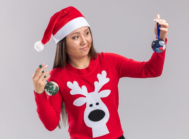 Thinking young asian girl wearing christmas hat with sweater holding and looking at christmas tree balls isolated on white background