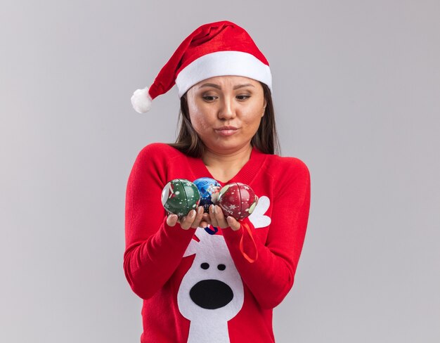 Думающая молодая азиатская девушка в новогодней шапке со свитером, держащая и смотрящую на елочные шары, изолированные на белом фоне
