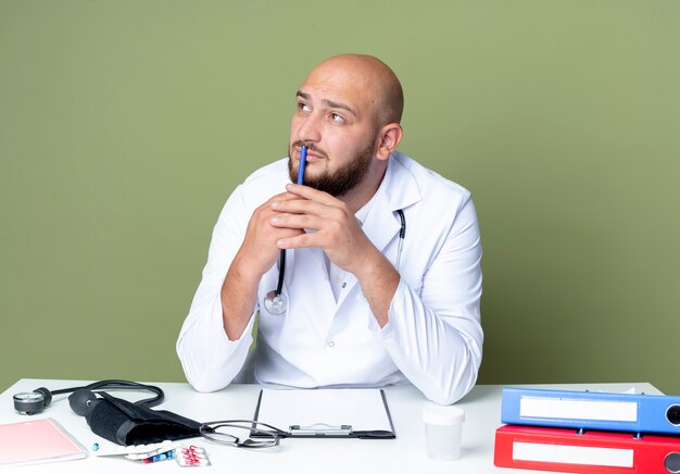 Thinking young bald male doctor wearing medical robe and stethoscope sitting at desk work with medical tools putting pen on mouth isolated on green wall