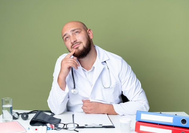 Думающий молодой лысый мужчина-врач в медицинском халате и стетоскопе сидит за столом
