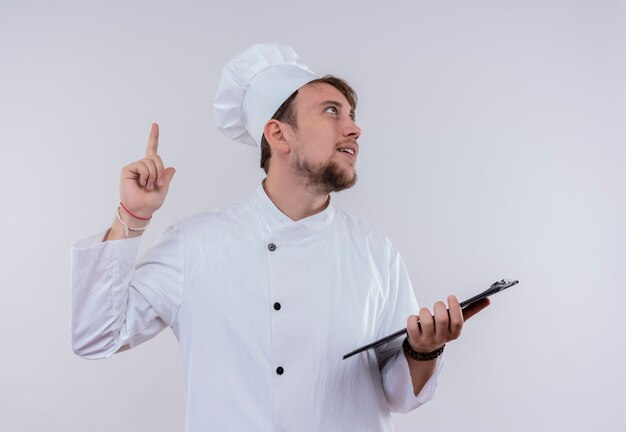 A thinking young bearded chef man wearing white cooker uniform and hat pointing up while holding blank folder on a white wall