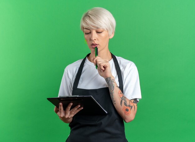 Thinking young beautiful female barber in uniform holding and looking at clipboard putting pen on chin isolated on green background