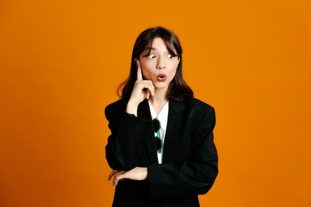 Thinking young beautiful female wearing black jacket isolated on orange background