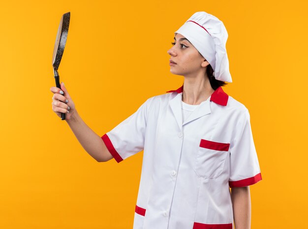Думая молодая красивая девушка в униформе шеф-повара, держащая и смотрящую на сковороду, изолированную на оранжевой стене