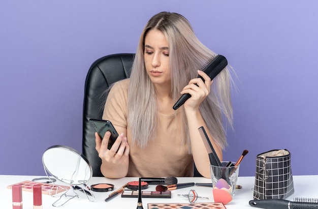 Думающая, молодая красивая девушка сидит за столом с инструментами для макияжа, расчесывая волосы, держа и глядя на телефон, изолированные на синем фоне