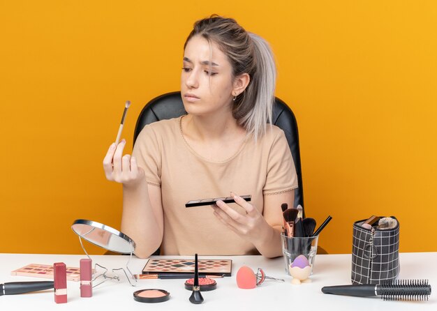 Thinking young beautiful girl sits at table with makeup tools holding and looking at eyeshadow palette with makeup brush isolated on orange background