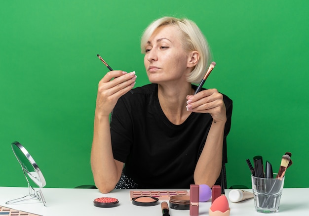 Thinking young beautiful girl sits at table with makeup tools holding and looking at makeup brushes isolated on green background