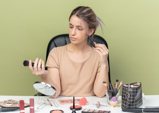 Thinking young beautiful girl sitting at desk with makeup tools holding and looking at makeup brush isolated on olive green background