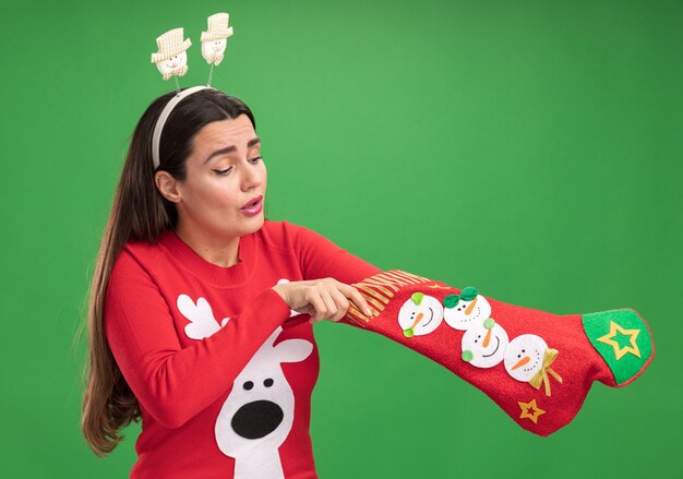 Thinking young beautiful girl wearing christmas sweater with christmas hair hoop putting hand in christmas socks isolated on green background