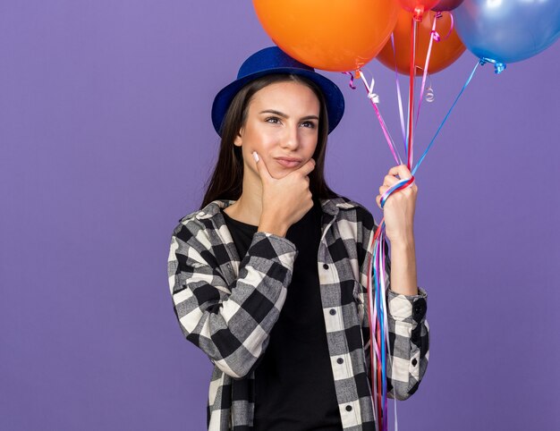 Pensando giovane bella ragazza che indossa cappello da festa tenendo palloncini afferrò il mento isolato sulla parete blu