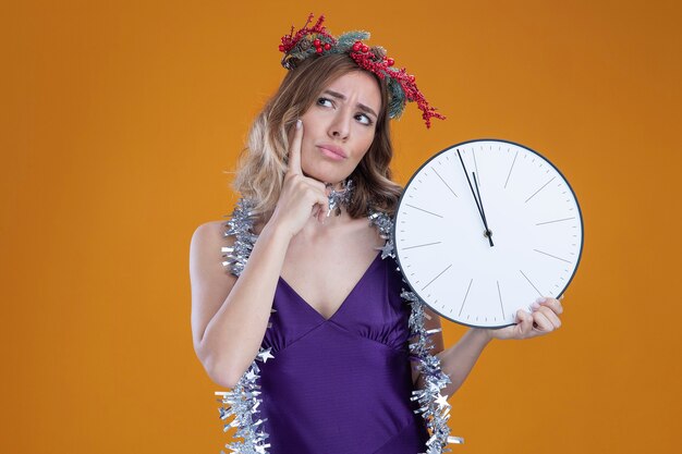 Thinking young beautiful girl wearing purple dress and wreath with garland on neck holding wall clock putting finger on cheek isolated on brown background