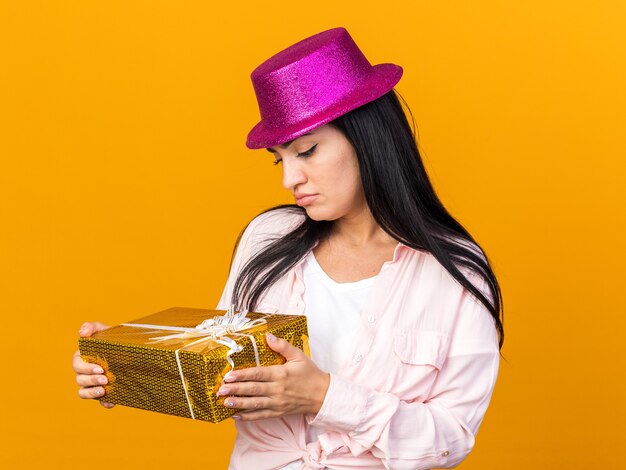 Thinking young beautiful woman wearing party hat holding and looking at gift box isolated on orange wall