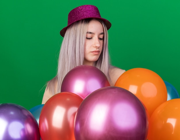 Thinking young beautiful woman wearing party hat standing behind balloons isolated on green wall