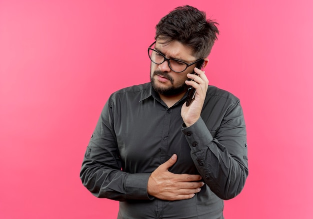 Thinking young businessman wearing glasses speaks on phone and putting hand on stomach isolated on pink