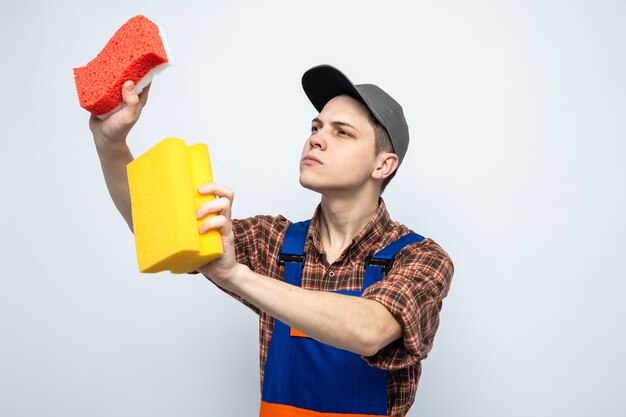 Thinking young cleaning guy wearing uniform and cap holding and looking at sponges 