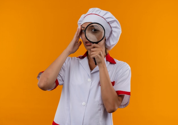 Thinking young cook female wearing chef uniform looking at camera with magnifier and putting hand on forehead on isolated orange background