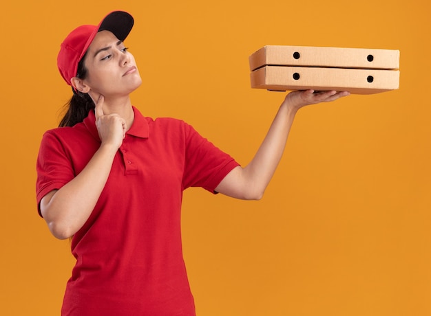 Thinking young delivery girl wearing uniform and cap holding and looking at pizza boxes isolated on orange wall