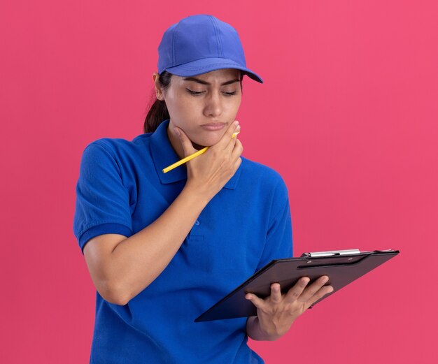 Thinking young delivery girl wearing uniform with cap holding and looking at clipboard grabbed chin isolated on pink wall