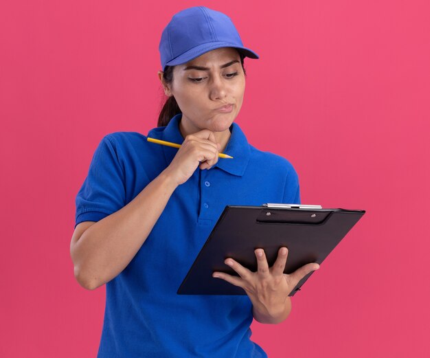 Thinking young delivery girl wearing uniform with cap holding and looking at clipboard putting hand on chin isolated on pink wall