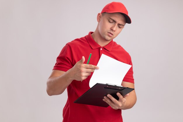 Thinking young delivery man wearing uniform with cap flipping through clipboard isolated on white wall